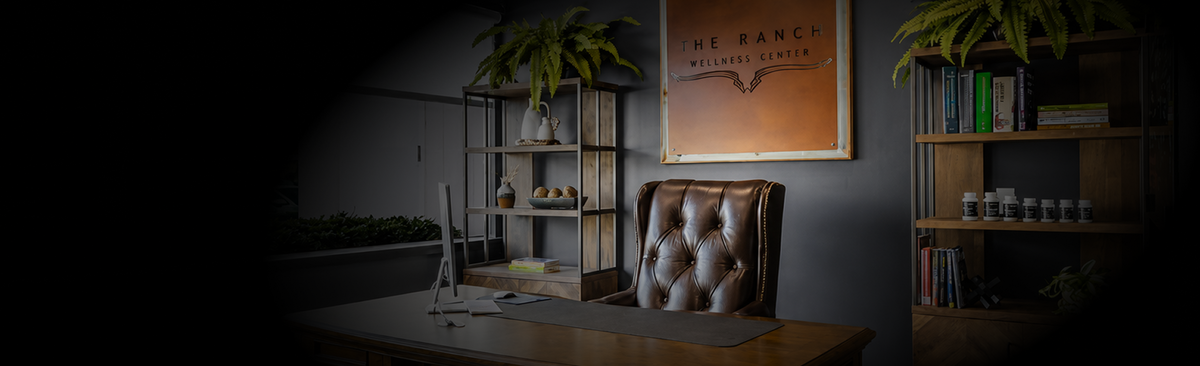 A dimly lit home office scene featuring a brown tufted leather office chair, a wooden desk, shelving units with books and plants, and a framed print on the wall with title THE RANCH WELLNESS CENTER.