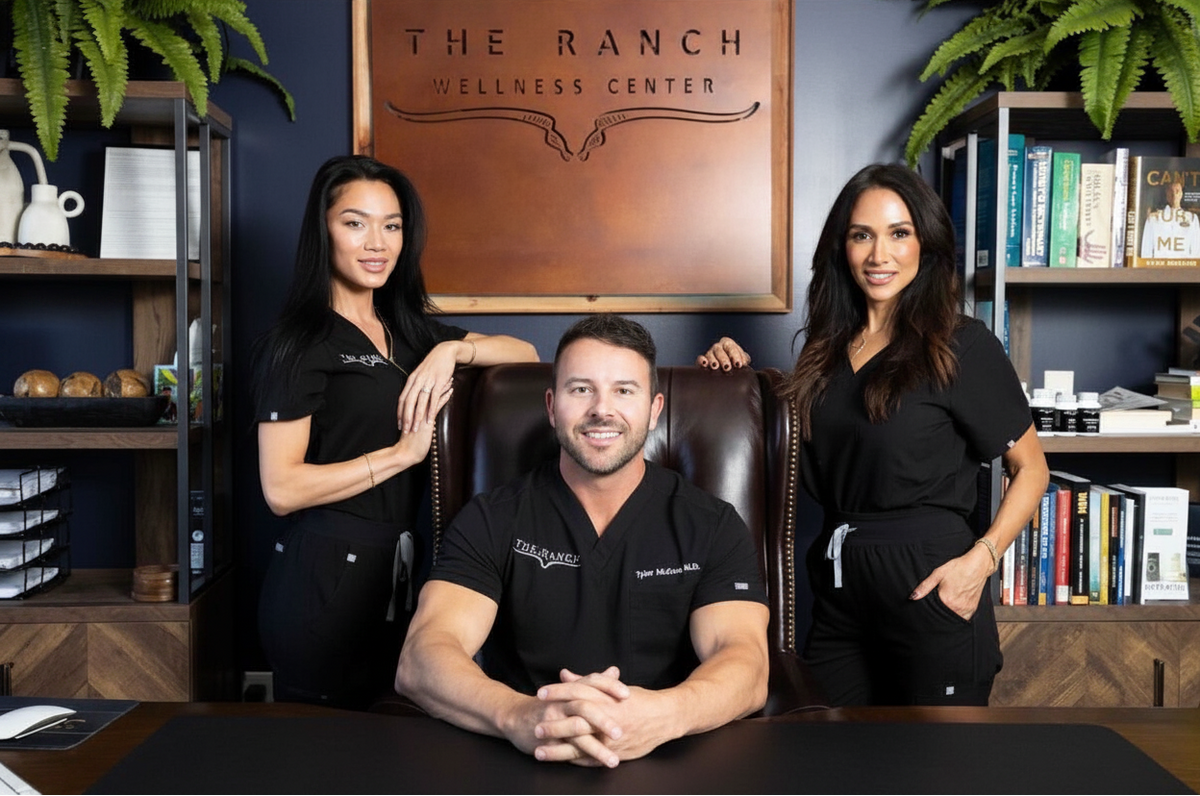 Three individuals in black attire standing behind a person in a chair with 'The Ranch Wellness Center' sign in the background.