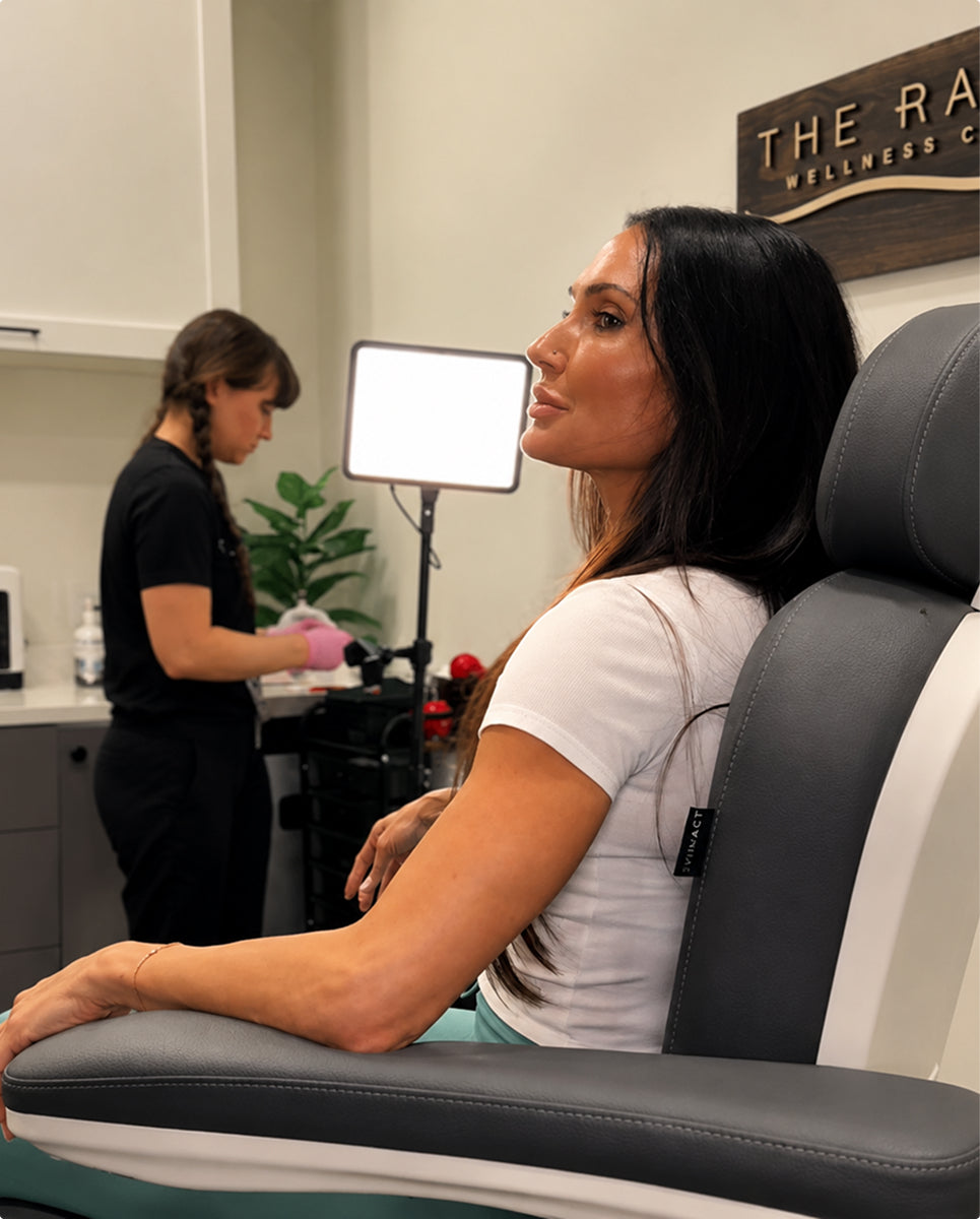 A woman with long dark hair sits in profile in a white and dark gray wellness chair inside a clinic. Another person in black stands in the background near a bright studio light, with a sign reading 'THE RA.