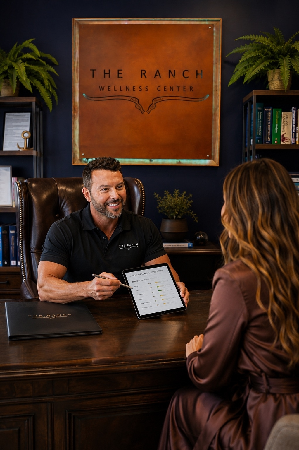 A man in a black polo shirt sits behind a wooden desk in an office, holding a tablet while talking to a woman in a brown robe. On the wall behind him is a large framed sign reading 'THE RANCH WELLNESS CENTER.