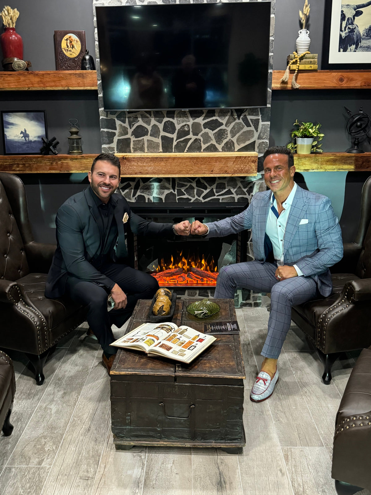 Two men in suits shaking hands in a room with a fireplace and wooden furniture.